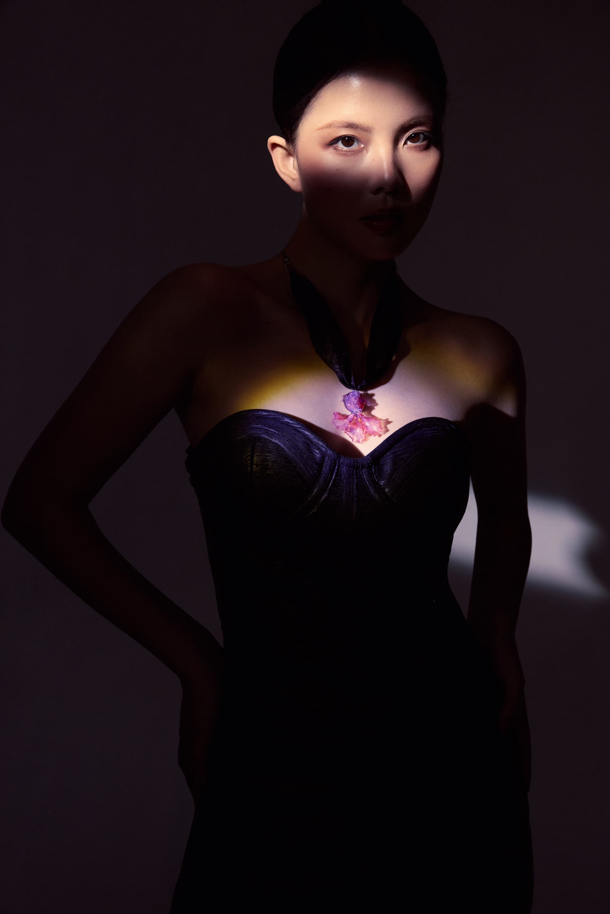 Model in black strapless dress with updo hair, hands on hips, gazing directly at camera — spotlight closeup on Koi Fish Pink Liuli Glass Choker Necklace and eyes against dark background