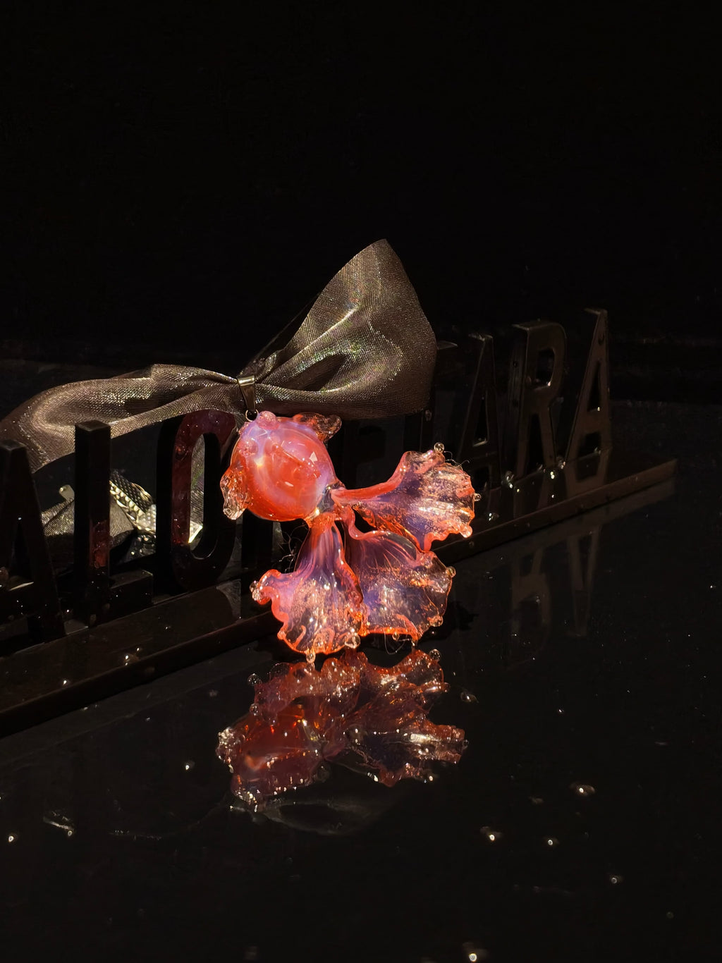 Close-up of handcrafted pink Liuli glass koi fish pendant, blush pink tones with crystal clear details, black organza ribbon, against black background