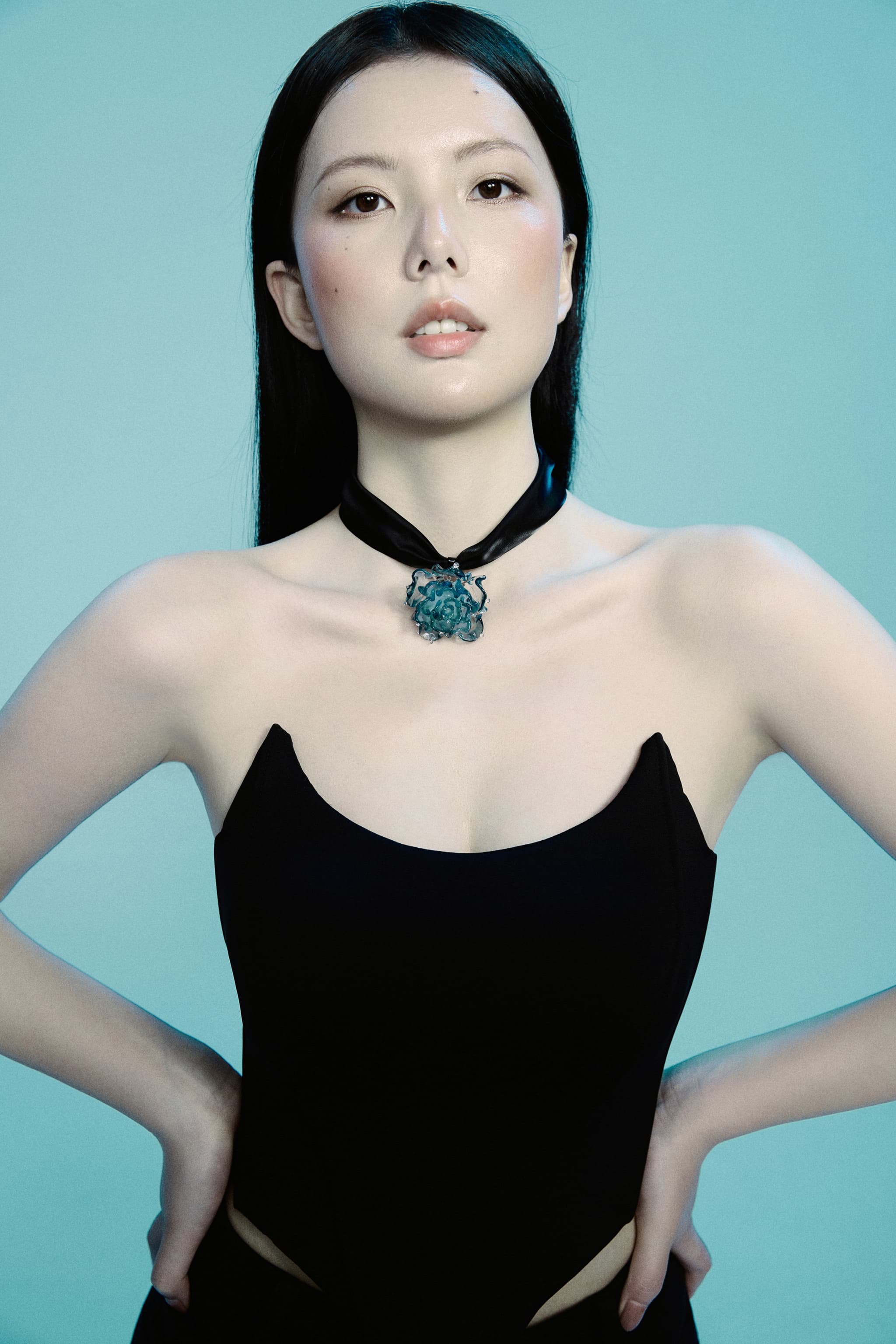 Model in black strapless top, hands on hips, chin slightly raised, gazing directly at camera wearing Ocean Free Blue Liuli Glass Rose Choker Necklace against solid blue background