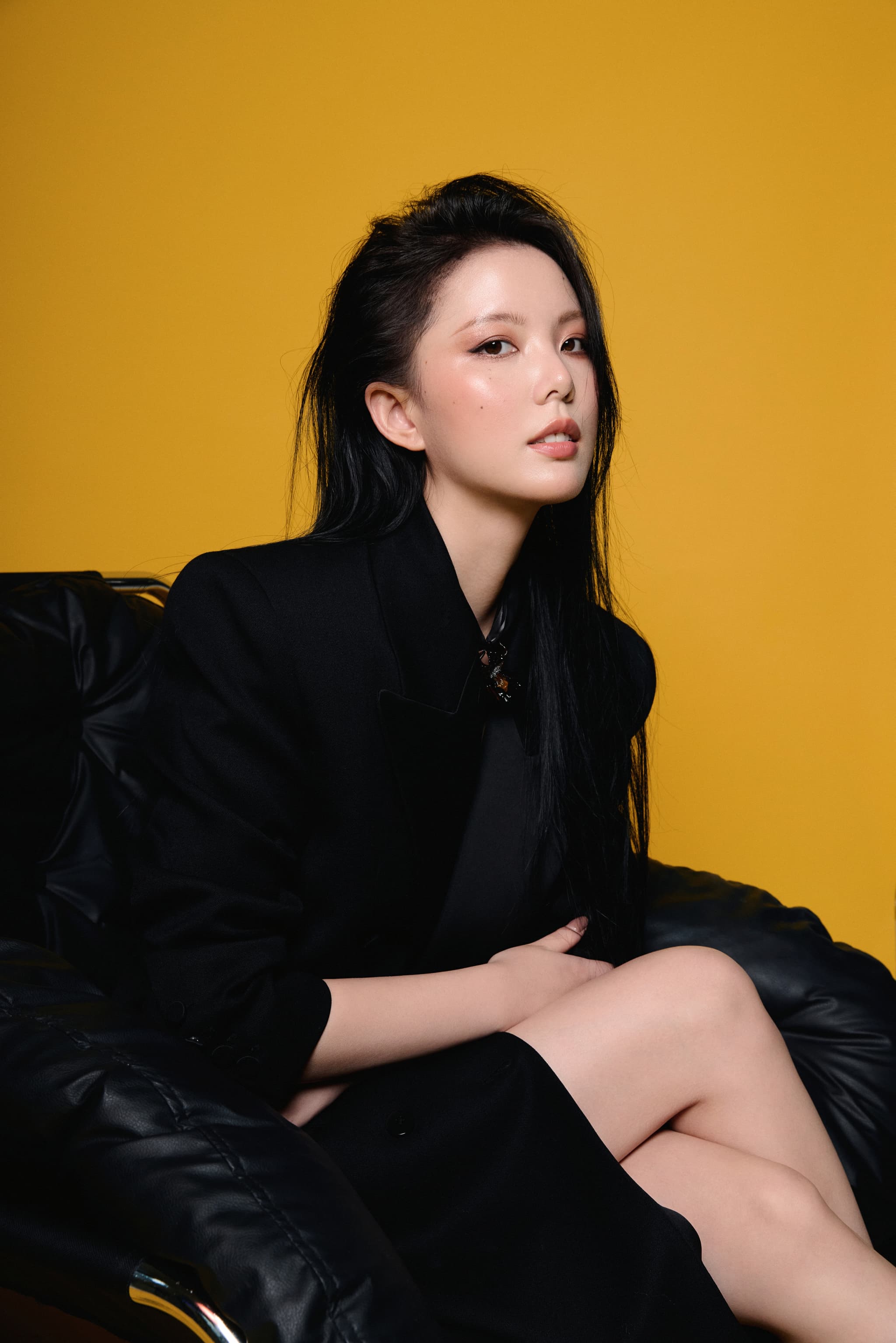 Model with straight black hair in black suit seated on black sofa, wearing Starfire Black Gold Liuli Glass Iris Choker Necklace, side profile gazing at camera against yellow background