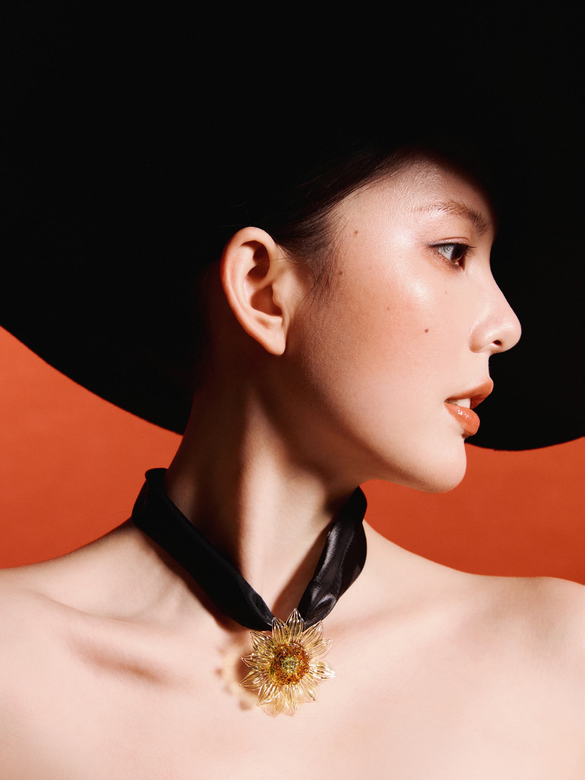 Model in black wide-brim hat and black strapless top wearing Youth Story Yellow Liuli Glass Sunflower Choker Necklace, side profile against red background
